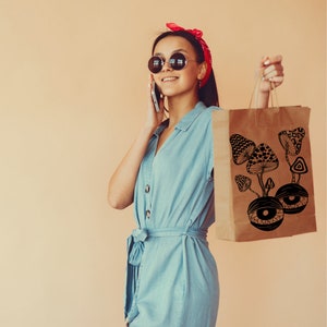 May include: A woman wearing a blue denim dress and sunglasses is holding a brown paper bag with a black and white illustration of mushrooms with eyes.
