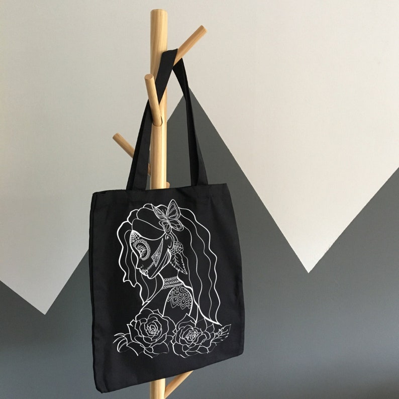 K&ouml;nnte beinhalten: Schwarze Tragetasche mit einem wei&szlig;en Design einer Frau mit einer Blumenkrone und filigranen Details. Das Design erinnert an einen Zucker Sch&auml;del.