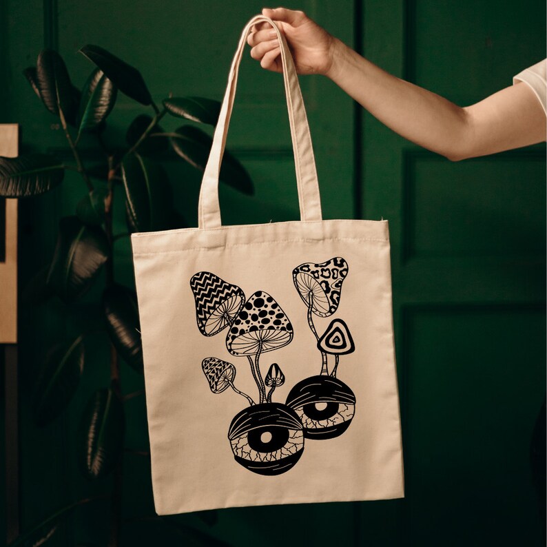 May include: A white canvas tote bag with black and white illustrations of mushrooms and eyes. The mushrooms have a psychedelic design and the eyes are large and round.