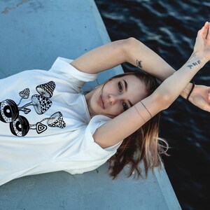 May include: A white t-shirt with a black and white graphic of mushrooms and eyes. The shirt is worn by a person lying on a gray surface.