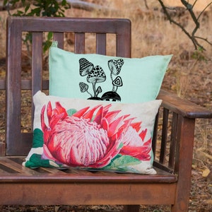 May include: Two throw pillows on a wooden bench. The top pillow is light green with a black and white illustration of mushrooms. The bottom pillow is white with a pink and green floral print.