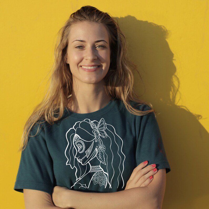 K&ouml;nnte beinhalten: Eine Frau mit langen blonden Haaren tr&auml;gt ein blaugr&uuml;nes T-Shirt mit einem wei&szlig;en Motiv einer Frau mit einem Schmetterling im Haar. Das Motiv ist eine Linienzeichnung mit filigranen Details.