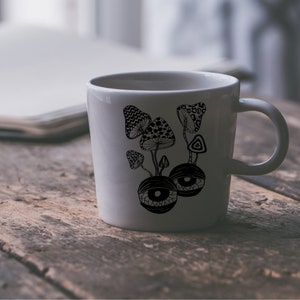 May include: White ceramic mug with a black and white illustration of mushrooms and eyes. The mushrooms are stylized and have intricate patterns. The eyes are large and have a psychedelic look.