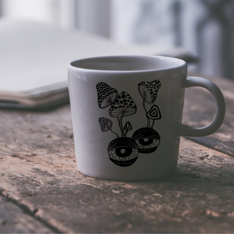 May include: White ceramic mug with a black and white illustration of mushrooms and eyes. The mushrooms are stylized and have intricate patterns. The eyes are large and have a psychedelic look.