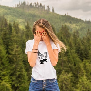 May include: A white t-shirt with a black and white graphic of mushrooms with eyes. The person wearing the shirt is covering their face with their hands.