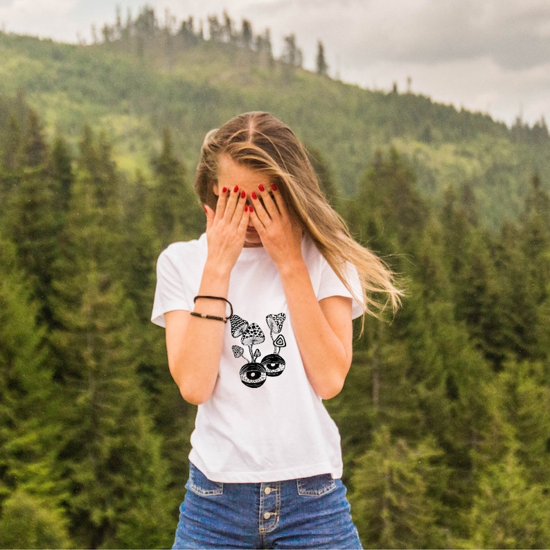 May include: A white t-shirt with a black and white graphic of mushrooms with eyes. The person wearing the shirt is covering their face with their hands.