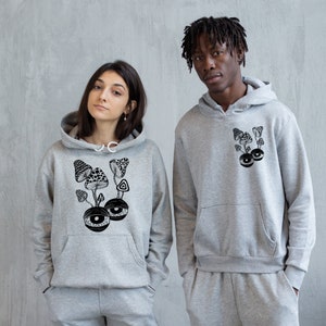 May include: Two grey hooded sweatshirts with a black and white graphic print of mushrooms and eyes. The graphic on the left sweatshirt features a cluster of mushrooms with eyes at the base. The graphic on the right sweatshirt features a cluster of mushrooms with a single eye at the base.