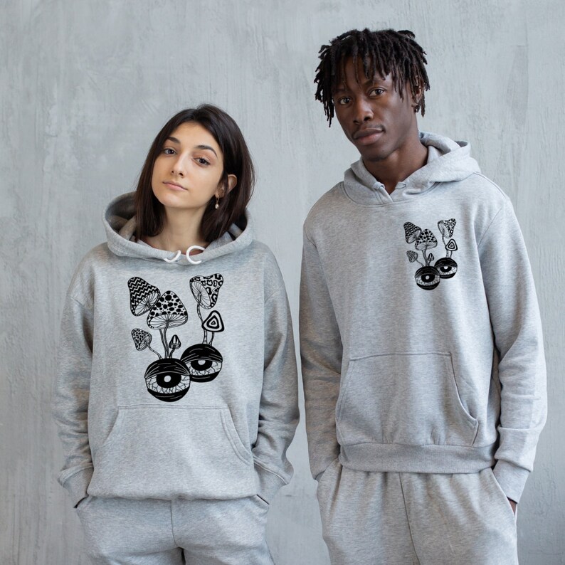 May include: Two grey hooded sweatshirts with a black and white graphic print of mushrooms and eyes. The graphic on the left sweatshirt features a cluster of mushrooms with eyes at the base. The graphic on the right sweatshirt features a cluster of mushrooms with a single eye at the base.