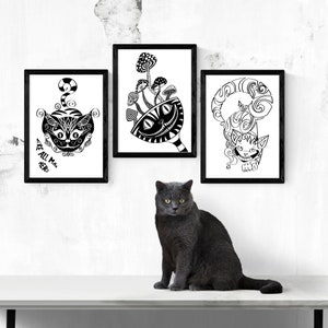 May include: Three framed black and white art prints featuring whimsical cat illustrations and mushroom designs. The text "WE'RE ALL MAD HERE" is visible on one print. A gray cat sits in front of the artwork.