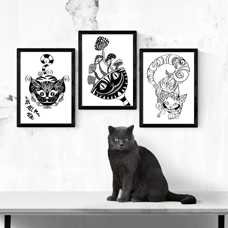 May include: Three framed black and white art prints featuring whimsical cat illustrations and mushroom designs. The text "WE'RE ALL MAD HERE" is visible on one print. A gray cat sits in front of the artwork.