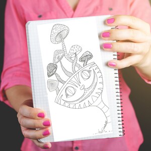 May include: A black and white illustration of the Cheshire Cat from Alice in Wonderland with mushrooms. The drawing is held in a spiral-bound notebook. The cat's grin and the mushrooms are detailed with intricate patterns.