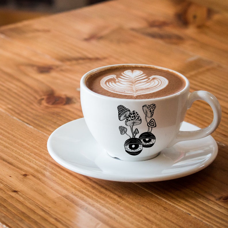 May include: A white ceramic coffee mug with a black and white design of mushrooms and eyes. The mug is filled with coffee and has a brown wooden table in the background.