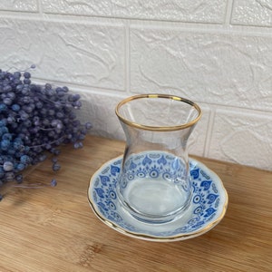 Puede incluir: Un platillo de cerámica blanco con un patrón azul y dorado que presenta un diseño de ojo turco. Una taza de té de vidrio transparente con un borde dorado se encuentra sobre el platillo.