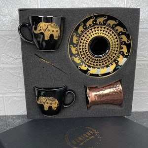 Op de afbeelding: Een zwarte cadeaudoos met een gouden inscriptie die "Unique" zegt en een gouden olifantontwerp. De doos bevat twee zwarte keramische koffiekopjes met gouden olifantontwerpen, een zwarte schotel met een gouden olifantontwerp en een koperen kopje.