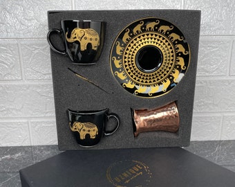 May include: A black gift box with a gold inscription that reads "Unique" and a gold elephant design. The box contains two black ceramic coffee cups with gold elephant designs, a black saucer with a gold elephant design, and a copper cup.