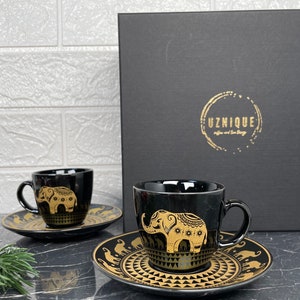 Op de afbeelding: Twee zwarte koffiekopjes met gouden olifantmotieven en bijpassende schoteltjes. De kopjes en schoteltjes hebben een gouden geometrisch patroon. De kopjes staan op een zwart oppervlak. De doos op de achtergrond is zwart met gouden letters die "Uznique Coffee and Tea Store" zeggen.