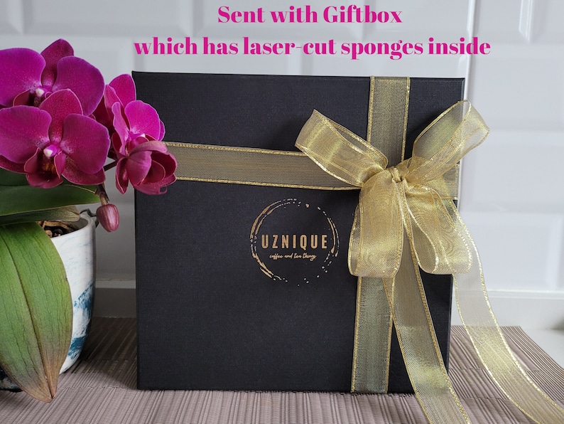 May include: A black gift box with a gold ribbon and bow. The box has a gold logo that says "UZNIQUE coffee and tea shop". The text "Sent with Giftbox which has laser-cut sponges inside" is written in pink above the box.