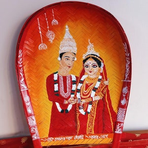 May include: A hand-painted bamboo tray depicting a traditional Indian wedding ceremony. The bride and groom are dressed in red and gold, with the groom wearing a white turban. The tray is decorated with intricate floral designs and the text "আবার আসা" in Bengali.