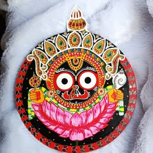 Op de afbeelding: Een ronde, handgeschilderde houten wanddecoratie met een godheid met grote witte ogen, een rood gezicht en een roze lotusbloem. Het ontwerp bevat ingewikkelde details met gouden, rode, groene en blauwe accenten. De achtergrond is zwart met een rode rand.