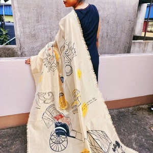 May include: A beige scarf with black and yellow illustrations of figures and vehicles. The scarf is draped over a person wearing a black dress. The illustrations include a rickshaw, a bridge, and other figures. The scarf has a fringed edge.