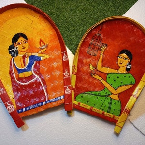 May include: Two hand-painted, woven bamboo fans. One fan features a woman in a red and blue sari holding a lamp against a yellow and orange background. The other fan depicts a woman in a green sari with a bird and cage against a red background.