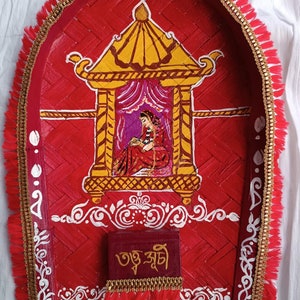 May include: A red, round, woven tray with a gold trim and fringe. The tray has a painted image of a woman in a yellow and gold palace with a pink curtain. The tray also has a small red box with gold trim and the text "তুই সুন্দরী" in Bengali.