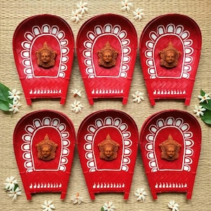 May include: Six red woven winnowing baskets arranged in two rows. Each basket features a white painted arch pattern and a central terracotta-colored relief sculpture of a deity face. Small white jasmine flowers are scattered around the baskets.
