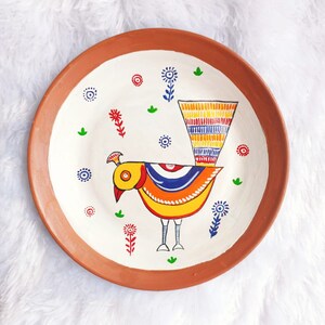 May include: A terracotta plate with a white background and a hand-painted design of a colourful bird with a long tail. The bird is surrounded by small flowers and leaves.