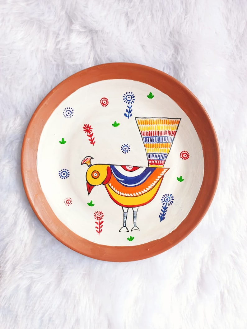 May include: A terracotta plate with a white background and a hand-painted design of a colourful bird with a long tail. The bird is surrounded by small flowers and leaves.