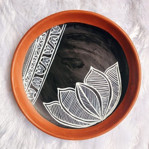 May include: A terracotta plate with a black background and white painted design. The design features a stylised floral pattern with intricate lines and curves.