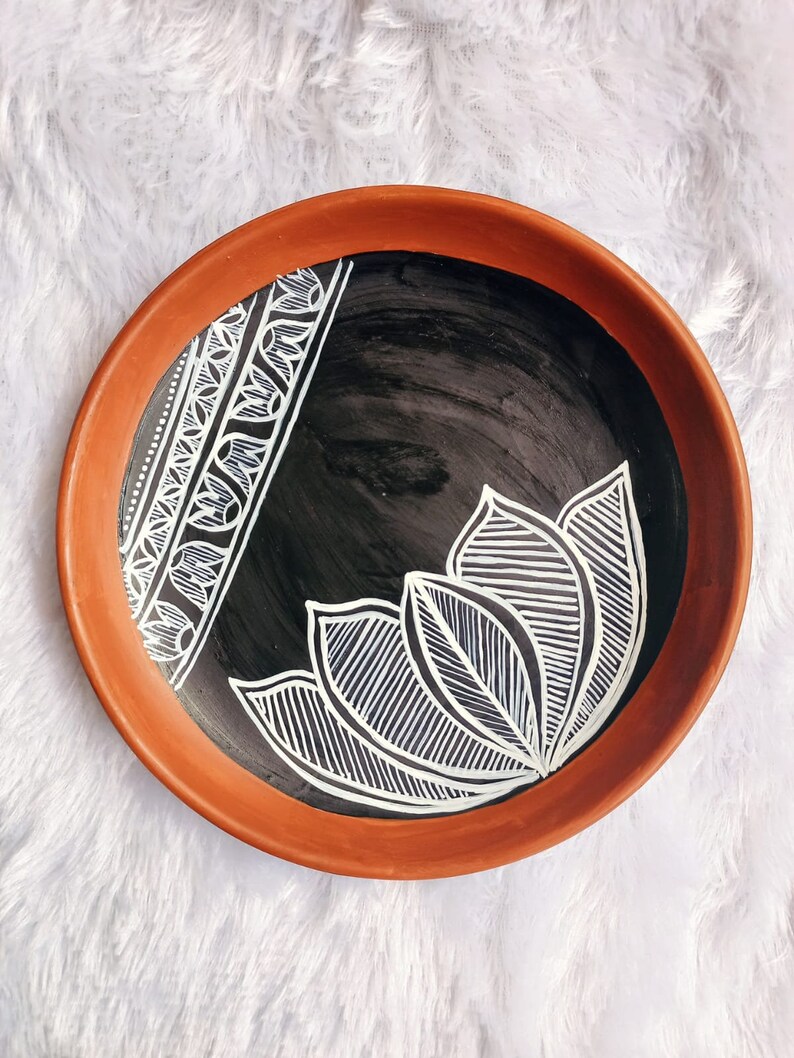 May include: A terracotta plate with a black background and white painted design. The design features a stylised floral pattern with intricate lines and curves.