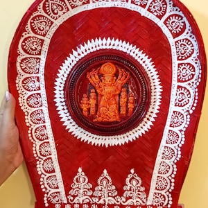 May include: A red woven wall hanging with white painted details. The design includes a central circular relief sculpture of a deity, surrounded by intricate patterns and a decorative border. The piece is held by hands.