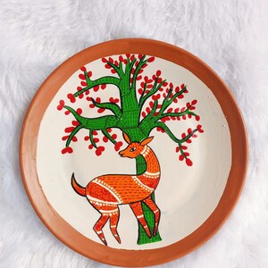 May include: A terracotta plate featuring a hand-painted design of a deer standing beneath a green tree with red blossoms. The deer is orange and white with intricate details.