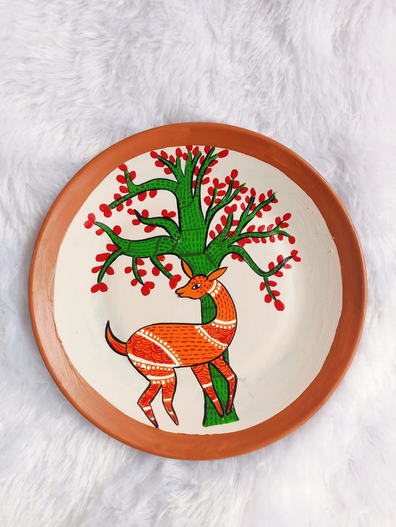 May include: A terracotta plate featuring a hand-painted design of a deer standing beneath a green tree with red blossoms. The deer is orange and white with intricate details.