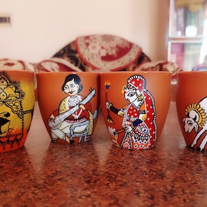May include: Four terracotta mugs with hand-painted designs. The mugs feature scenes of people playing music, a woman in traditional clothing, and a woman milking a cow. The mugs are a vibrant orange color with black, white, yellow, red, and green accents.