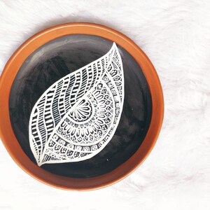 May include: A terracotta plate with a black centre and a white mandala design in the shape of a leaf.