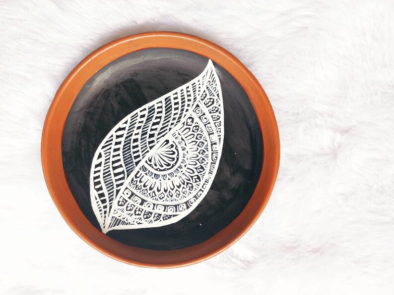 May include: A terracotta plate with a black centre and a white mandala design in the shape of a leaf.