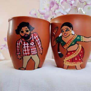 May include: Two terracotta-colored ceramic mugs with hand-painted figures. One mug features a man in a red and white plaid shirt and sunglasses. The other mug depicts a woman in a green and red sari, with gold jewelry.