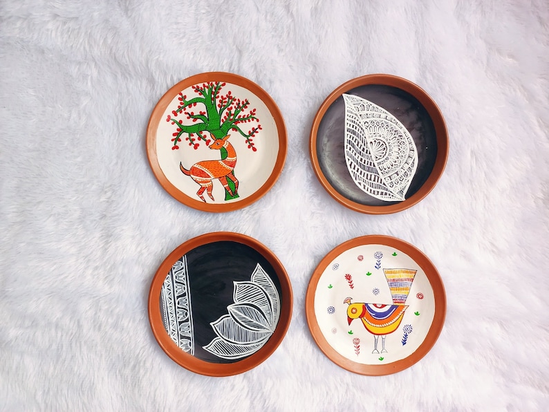 May include: Four terracotta plates with hand-painted designs. One plate features a deer under a tree, another has a stylised leaf, a third has a bird with a colourful tail, and the last has a floral design.