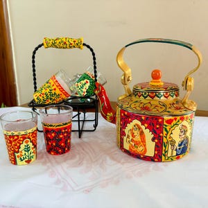 May include: A decorative tea set with a hand-painted kettle and matching glasses. The kettle is yellow, red, and gold with intricate designs and illustrations. The glasses are decorated with similar patterns. A black metal stand holds additional glasses.