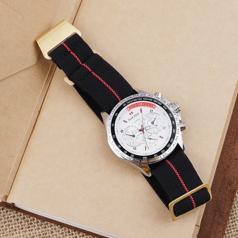 Puede incluir: Una correa de reloj negra y roja con una hebilla dorada. La esfera del reloj es blanca con marcas negras y el texto "NAKZEN" en rojo.