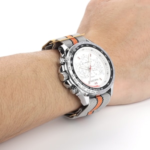Puede incluir: Un reloj de pulsera cron&oacute;grafo plateado con una esfera blanca y una banda de nylon a rayas naranja y gris. La esfera del reloj presenta una ventana de fecha, tres subesferas y el texto "NAKZEN".