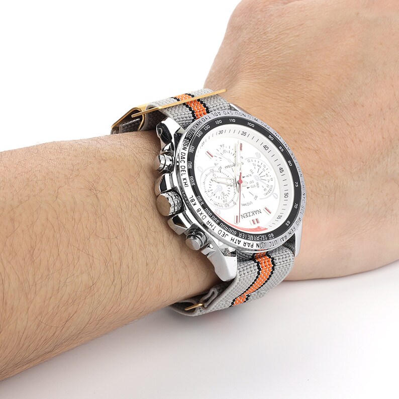 Puede incluir: Un reloj de pulsera cron&oacute;grafo plateado con una esfera blanca y una banda de nylon a rayas naranja y gris. La esfera del reloj presenta una ventana de fecha, tres subesferas y el texto "NAKZEN".