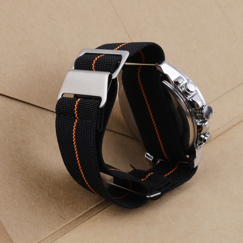Puede incluir: Una correa de reloj negra con costuras naranjas y una hebilla plateada. La correa est&aacute; hecha de nylon y est&aacute; dise&ntilde;ada para un reloj con una anchura de leng&uuml;eta de 20 mm.