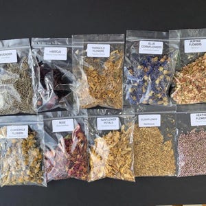 May include: An assortment of dried flowers in clear plastic bags, including lavender, hibiscus, marigold, blue cornflowers, jasmine, chamomile, rose, sunflower petals, elderflower, and heather flowers. Each bag has a label with the flower's name.
