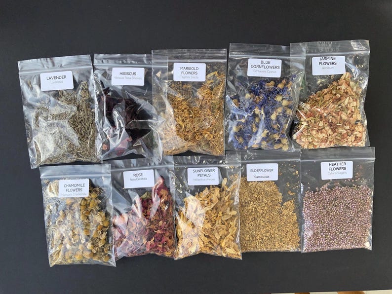 May include: An assortment of dried flowers in clear plastic bags, including lavender, hibiscus, marigold, blue cornflowers, jasmine, chamomile, rose, sunflower petals, elderflower, and heather flowers. Each bag has a label with the flower's name.
