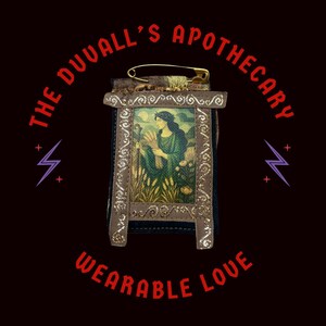 Può includere: Una piccola immagine incorniciata di una donna che tiene un fascio di grano. La cornice è in pelle marrone e ha un bordo dorato. L'immagine è fissata a uno spillo di sicurezza, rendendola indossabile. Il testo "THE DUVAL'S APOTHECARY WEARABLE LOVE" circonda l'immagine.