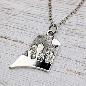 May include: A silver-toned pendant necklace featuring a trapezoidal charm with a textured mountain landscape design and a circular moon. The pendant hangs from a delicate silver-toned chain link, showcasing a polished finish on the lower section.