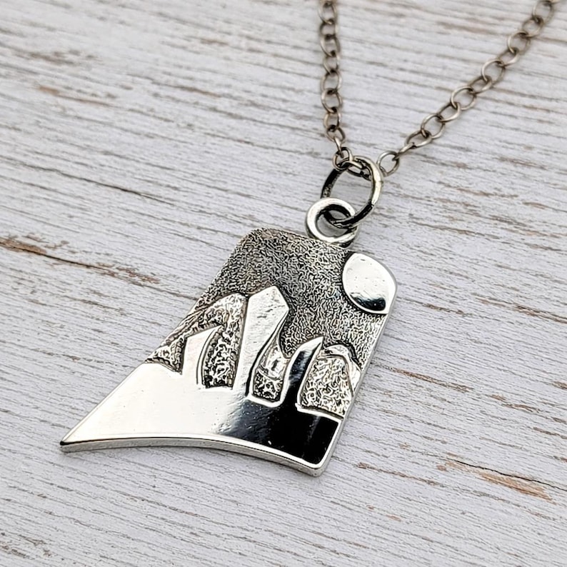 May include: A silver-toned pendant necklace featuring a trapezoidal charm with a textured mountain landscape design and a circular moon. The pendant hangs from a delicate silver-toned chain link, showcasing a polished finish on the lower section.