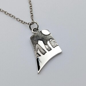 May include: A silver-toned pendant necklace featuring a trapezoidal charm with a textured, dark-finished mountain range and a circular moon design. The pendant hangs from a delicate silver-toned chain with round links, showcasing a polished finish.
